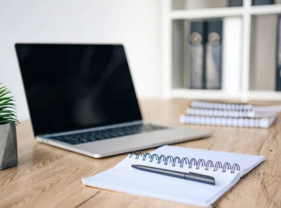 A slim budget laptop under $500 on the working desk; with pen, note books and a mini pot of plant next to it